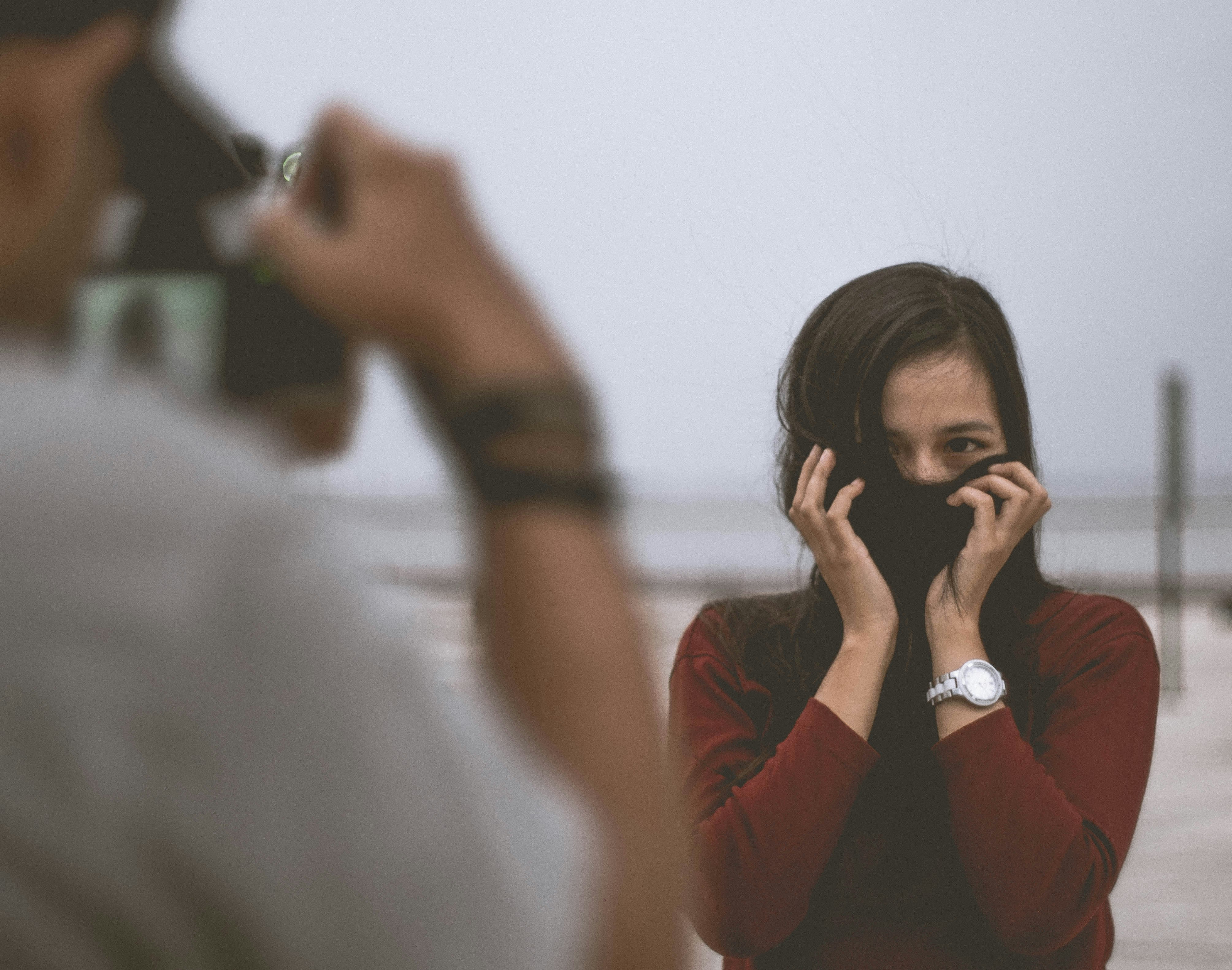 selective focus photography of person wearing white shirt taking photo of woman wearing red long-sleeved shirt covering her face with hair